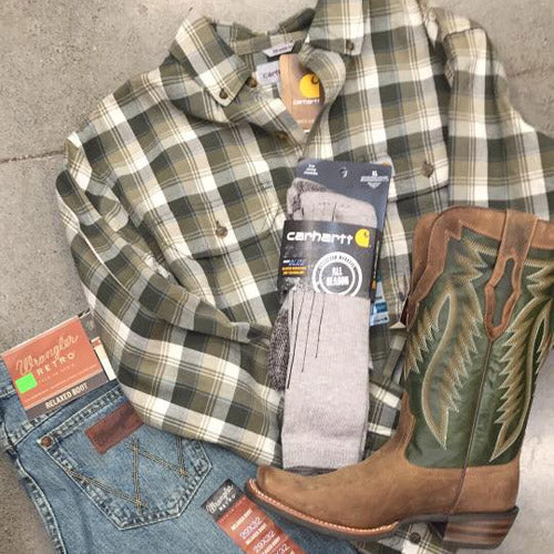 Plaid flannel shirt, jeans, and Carhartt socks lay on a floor next to a pair of brown cowboy boots with green accents, showcasing a rustic style.