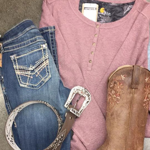 A laid-back country outfit with a pink henley shirt, distressed blue jeans, a studded leather belt, and rustic cowboy boots on a gray surface.