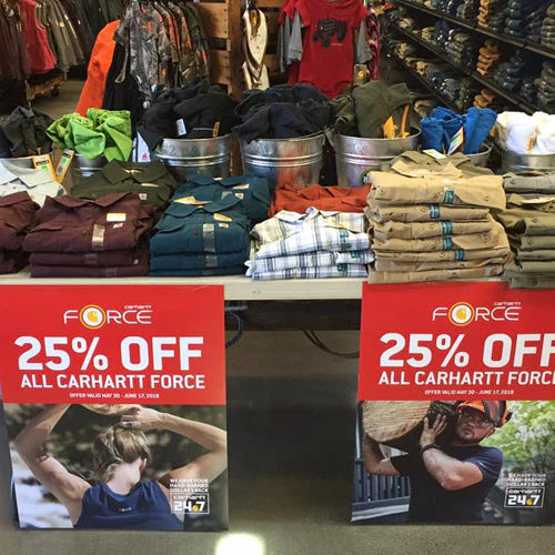 Retail display with folded Carhartt clothing on a table, surrounded by metal bins. Two red signs advertise a 25% discount on Carhartt Force apparel.