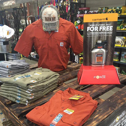 Display of outdoor work shirts in a store, featuring an orange button-up shirt on a mannequin, stacked shirts, and a free canteen promotion.