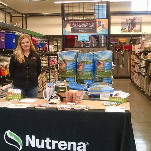 A woman stands by a Nutrena display table in a store aisle. The table is covered with promotional material, next to stacked bags of animal feed.